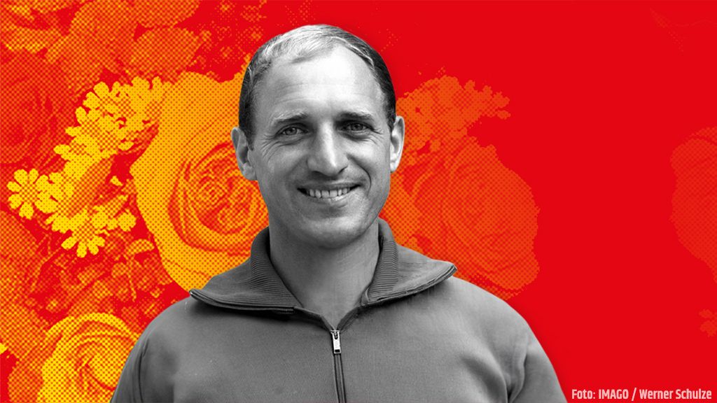 Mann mit kurzem Haar lächelt vor einem roten Hintergrund mit Blumenmotiven. Er trägt einen Reißverschluss-Pullover.