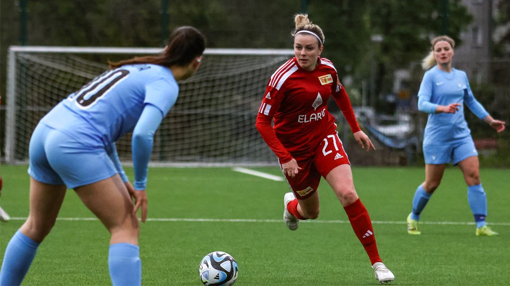 Eine Fußballspielerin in roter Uniform dribbelt den Ball auf einem Kunstrasenplatz, während eine Spielerin in blauer Uniform sie verfolgt.