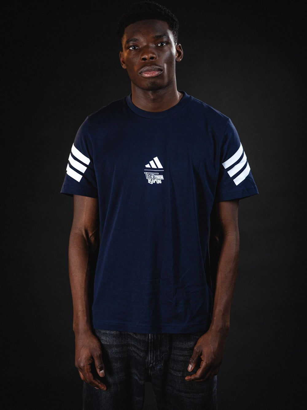 Ein junger Mann trägt ein dunkelblaues T-Shirt mit weißen Streifen an den Ärmeln vor einem dunklen Hintergrund.