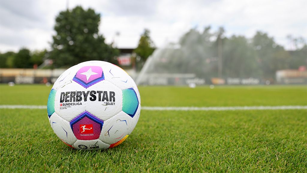 A football with the inscription "DERBYSTAR" lies on a green grass field, with a sprinkler system visible in the background.