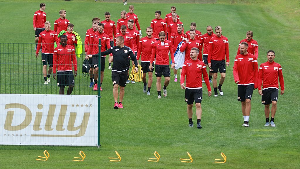 Eine Gruppe von Athleten in roten Trainingsanzügen geht auf einem grünen Rasenplatz, während Trainingsutensilien im Vordergrund liegen.