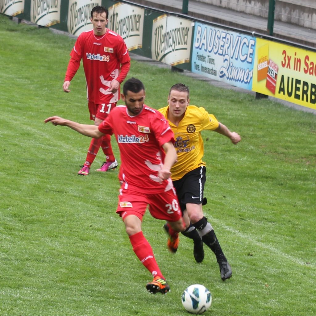 Zwei Fußballspieler im Wettkampf um den Ball auf einem grünen Spielfeld, während ein weiterer Spieler im Hintergrund zusieht.