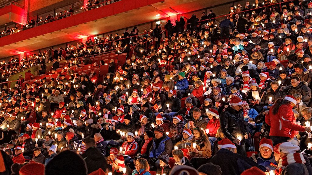 A large crowd in festive clothing wearing Christmas hats is holding candles in the dark.
