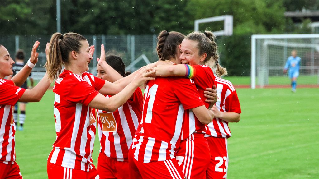 Players of the football team in red jerseys celebrate a goal on the field, surrounded by cheering teammates.