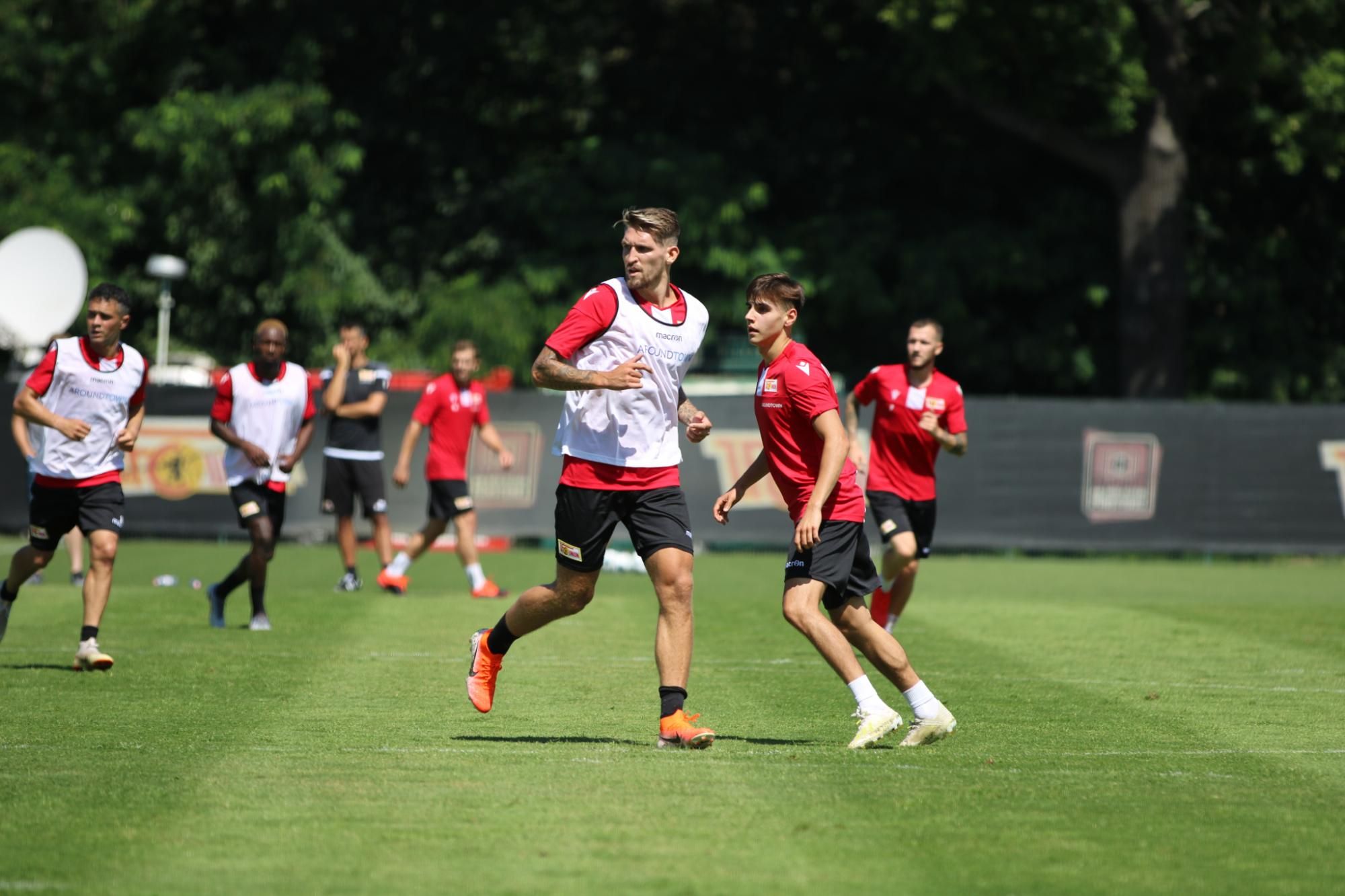 Zwei Fußballspieler in Trainingstrikots auf einem grünen Sportplatz, umgeben von weiteren Spielern im Hintergrund.