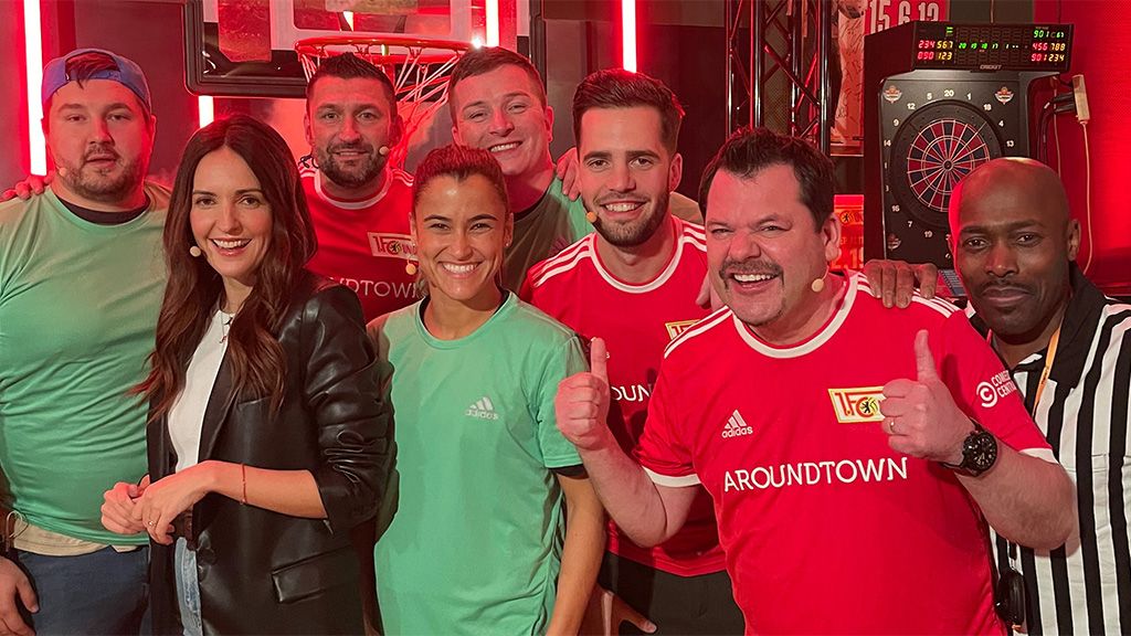 Eine Gruppe von acht Personen steht lächelnd zusammen, einige tragen Fußballtrikots, andere T-Shirts. Im Hintergrund sind Sportgeräte sichtbar.