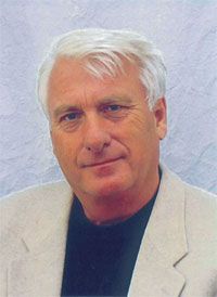 Porträt eines älteren Mannes mit blondem Haar, in einem hellen Blazer und dunklem Oberteil, vor einem neutralen Hintergrund.