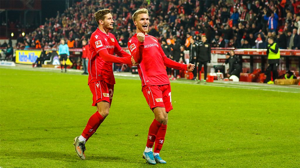 Zwei Fußballspieler in roten Trikots feiern jubelnd auf dem Spielfeld vor einer jubelnden Menge.