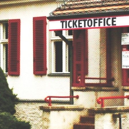 Ein Ticketbüro mit rotem Schild, großen Fenstern und roten Fensterläden, umgeben von einer kleinen Hecke.