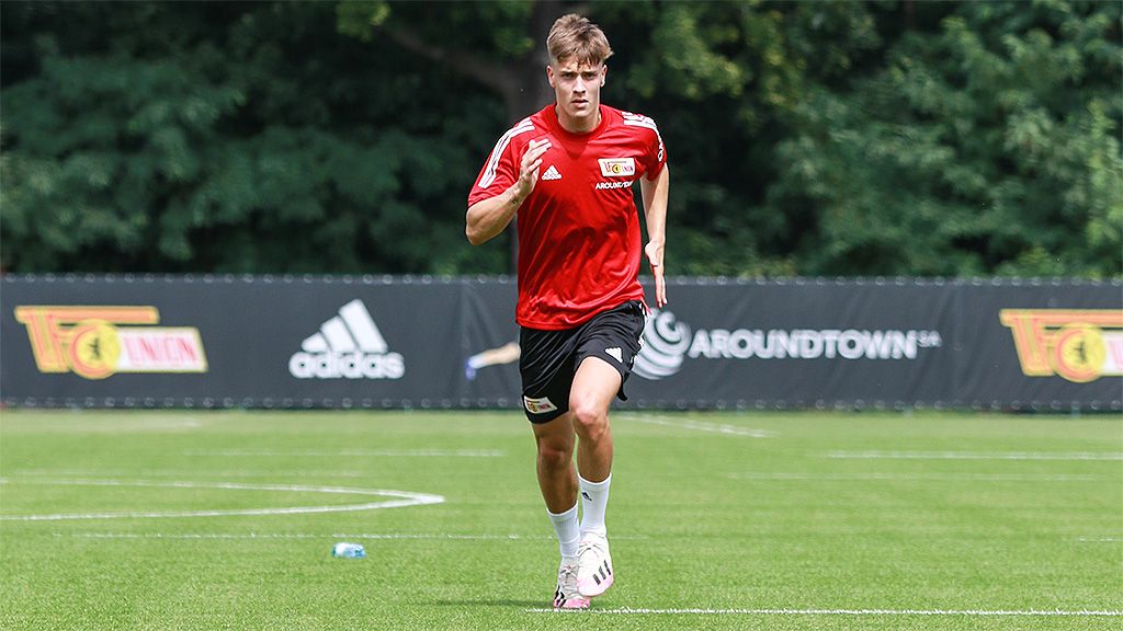 A young man in a red jersey is jogging on a soccer field with green grass and an advertising banner in the background.