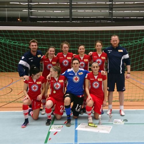 Eine Gruppe von weiblichen Fußballspielern in roten Trikots mit einem Trainer, aufgestellt vor einem Fußballfeld.