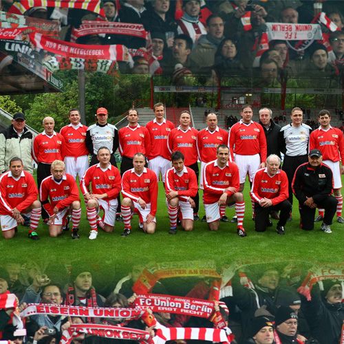 Eine Gruppe von Fußballspielern in roten Trikots steht auf einem Platz, umgeben von Fans mit Schals des FC Union Berlin.