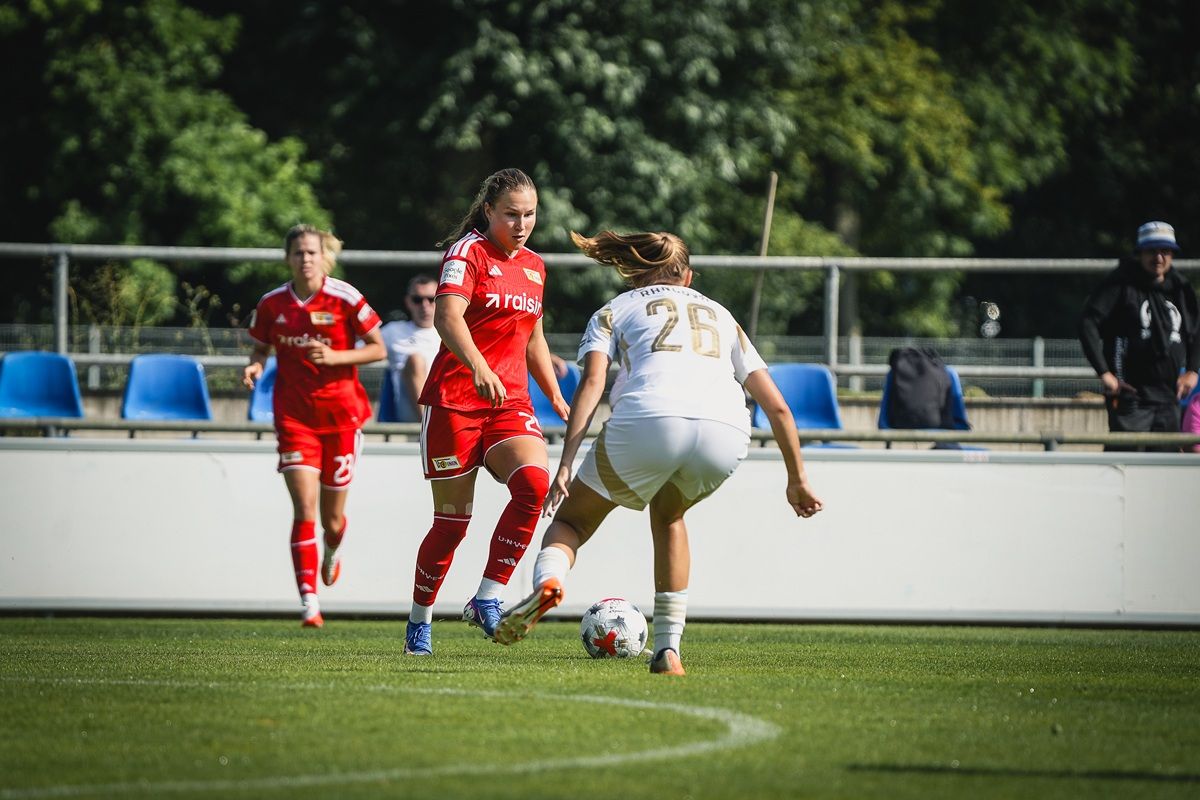 Zwei Frauenfußballspielerinnen stehen auf einem Spielfeld, während eine dritte Spielerin den Ball dribbelt. Zuschauer im Hintergrund.