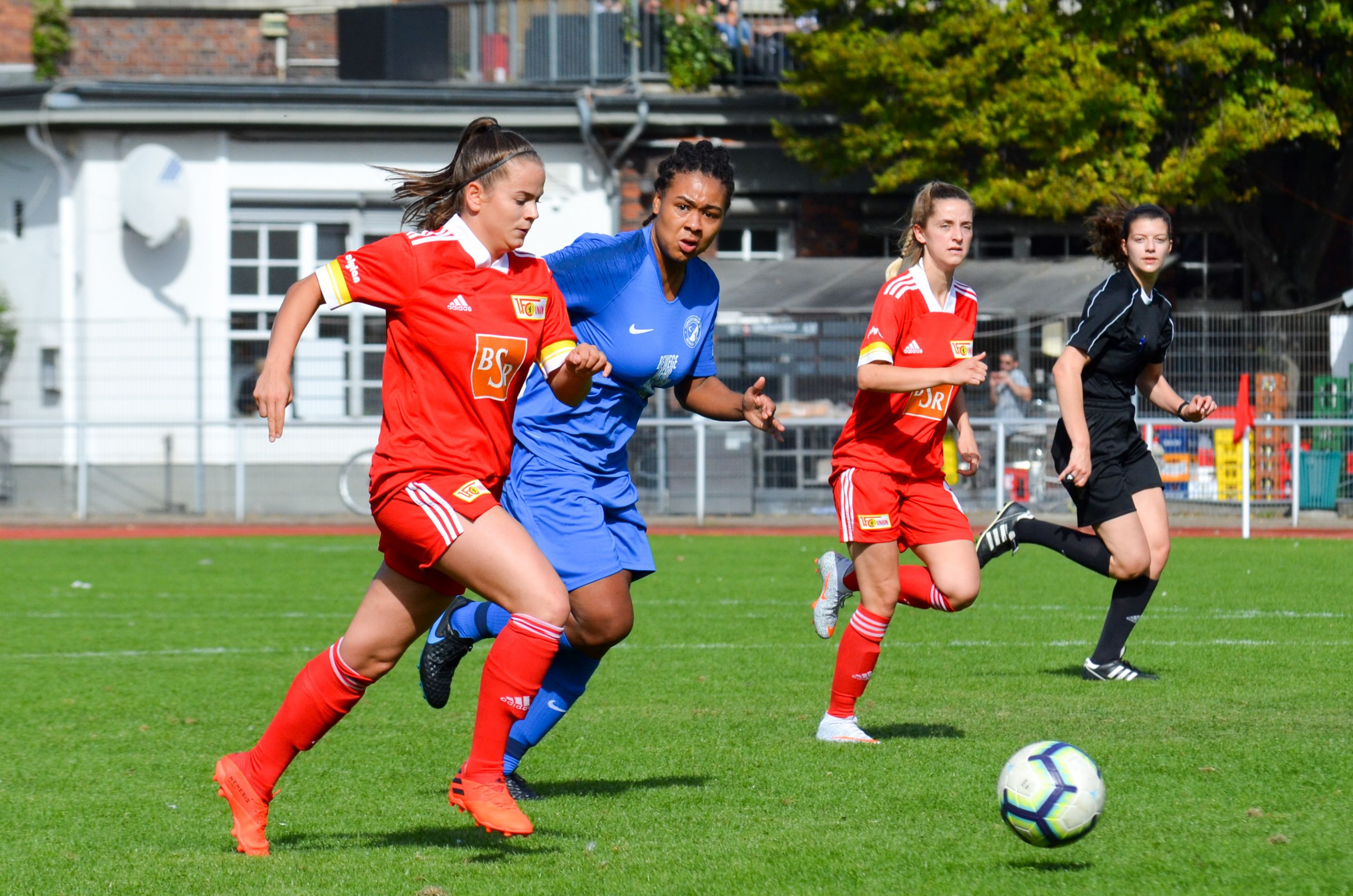 Drei Fußballspielerinnen im Wettlauf, eine in blauer, zwei in roter Uniform, auf einem grünen Spielfeld mit einem Schiedsrichter im Hintergrund.