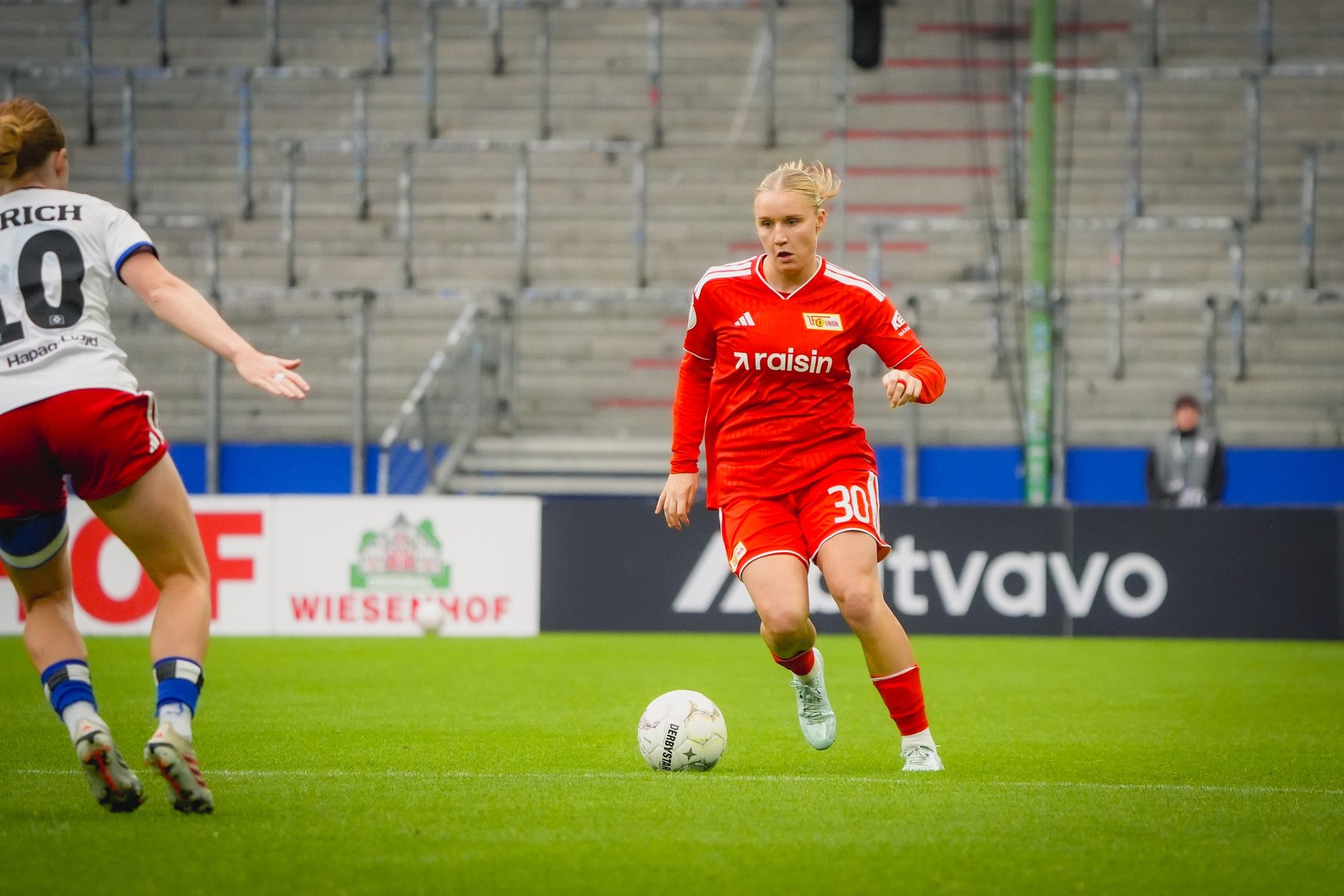 Fußballspielerin in rotem Trikot dribbelt den Ball auf dem Spielfeld, während eine Gegenspielerin in weißem Trikot näher kommt.