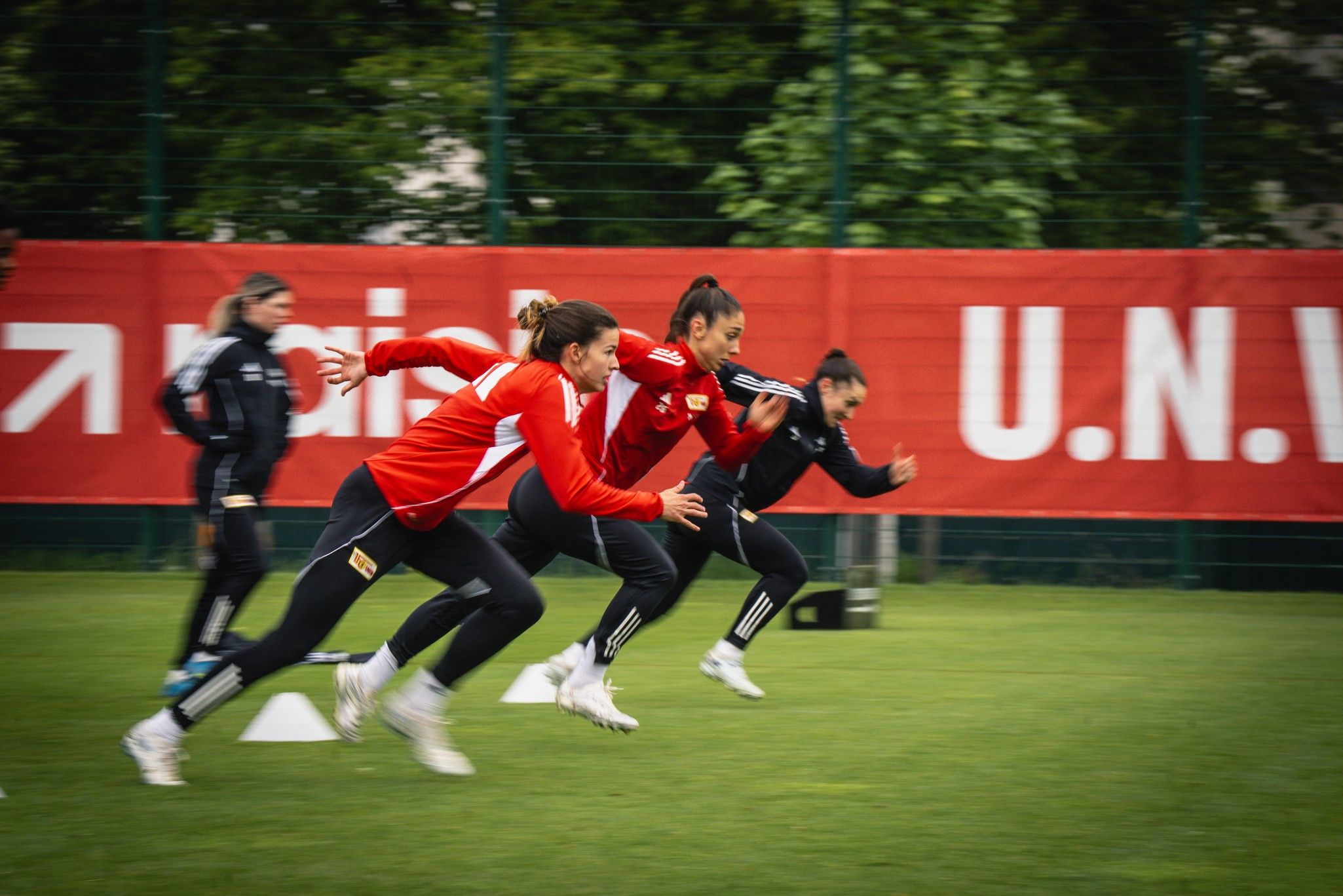 Drei Frauen in roten Trikots sprinten auf einem Fußballfeld während des Trainings. Hintergrund mit Werbebannern.