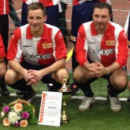 Zwei Sportler in roten und weißen Trikots posieren mit einem Pokal und einer Urkunde, umgeben von Blumen.