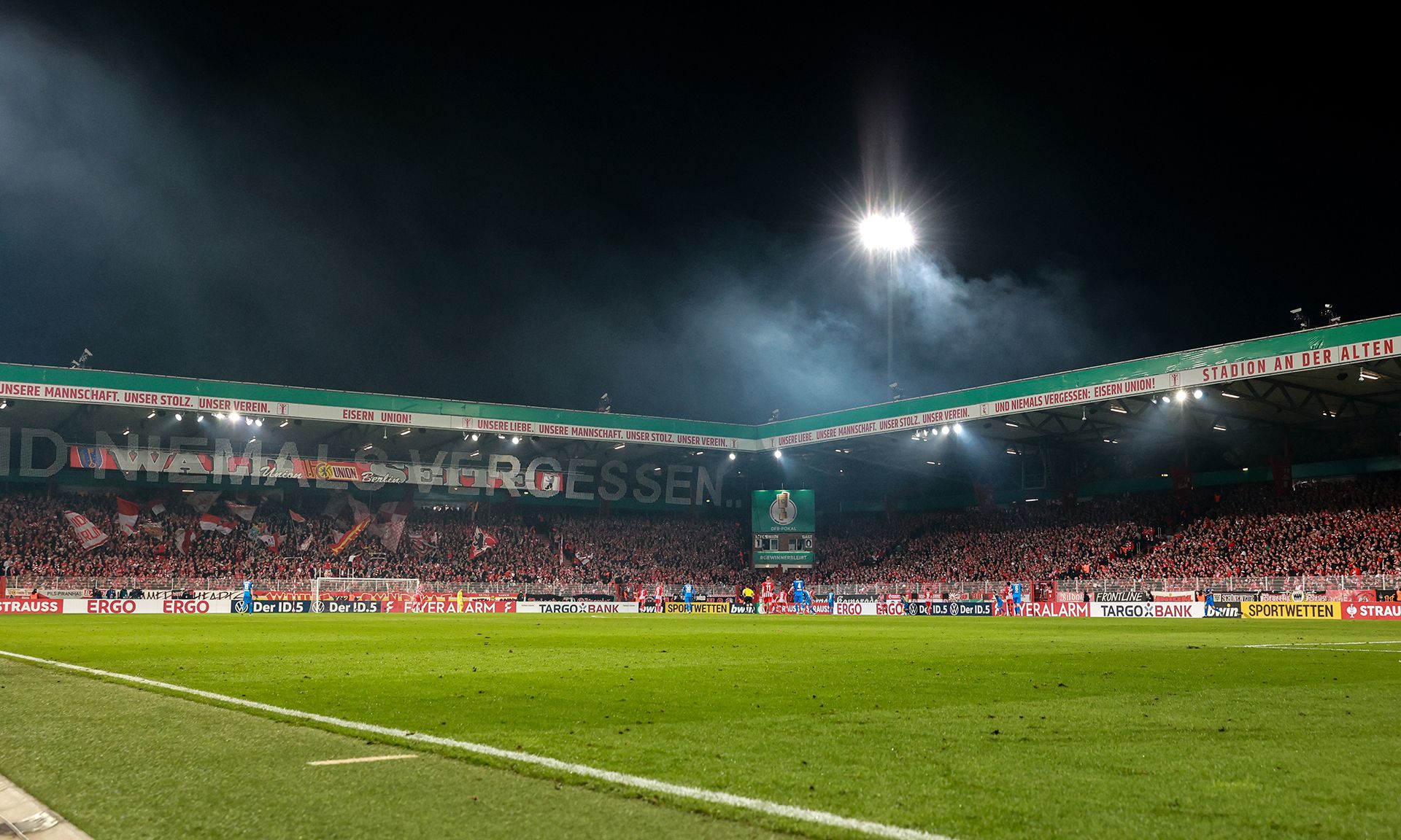 Blick auf ein vollbesetztes Fußballstadion bei Nacht, umgeben von festlicher Beleuchtung und rauchenden Effekten.