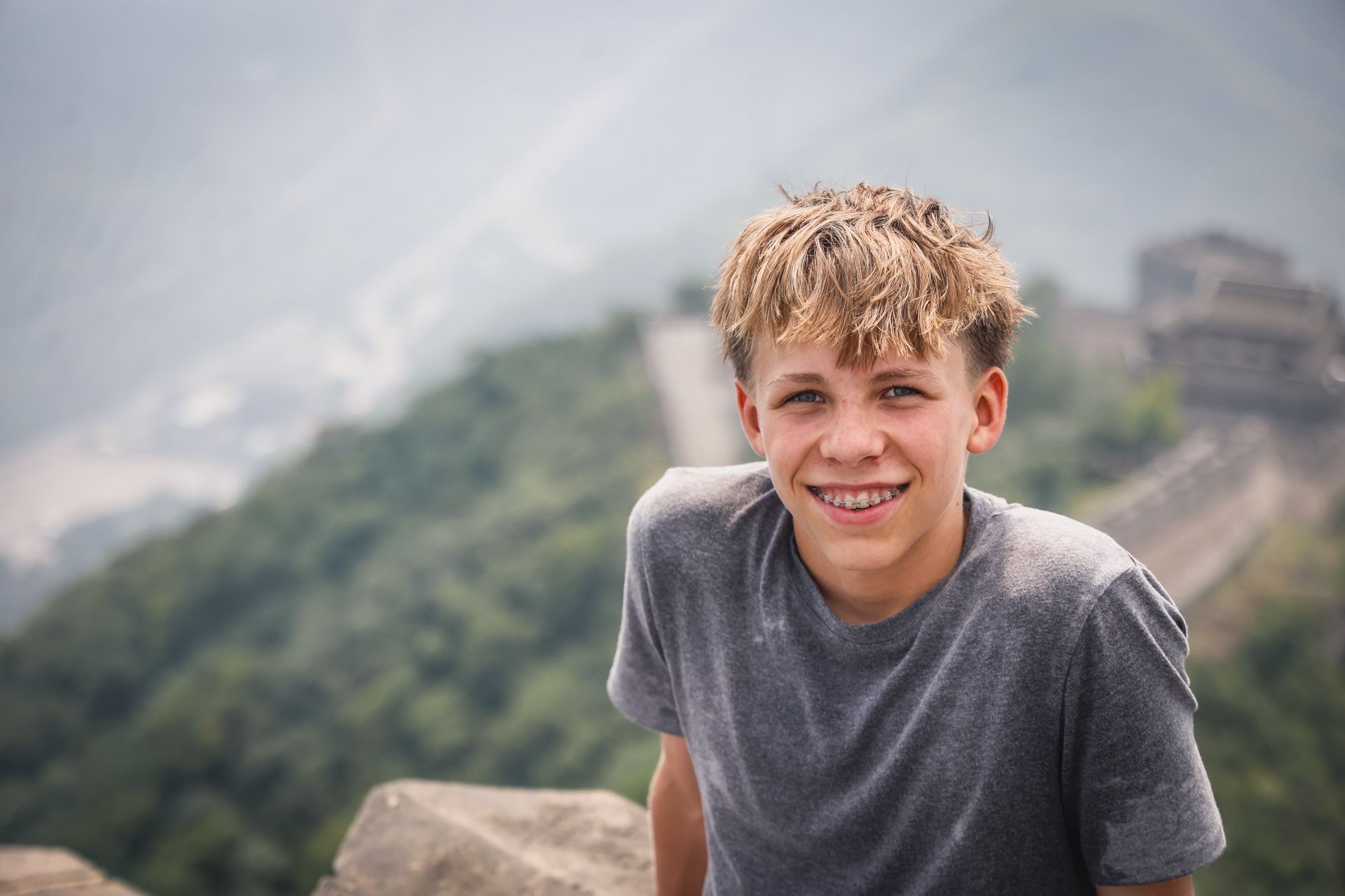 Junger Mann mit blonden, kurzen Haaren lächelt vor einer hügeligen Landschaft mit einer Struktur im Hintergrund.
