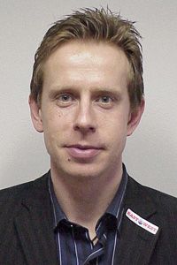 Ein Mann mit blonden Haaren und einem Holzfällerhemd, der in einem grauen Anzug posiert und leicht in die Kamera lächelt.