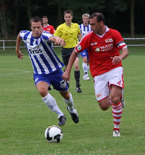Zwei Fußballspieler in Aktion auf dem Rasen, ein blauer und ein roter Trikot kämpfend um den Ball. Zuschauer im Hintergrund.