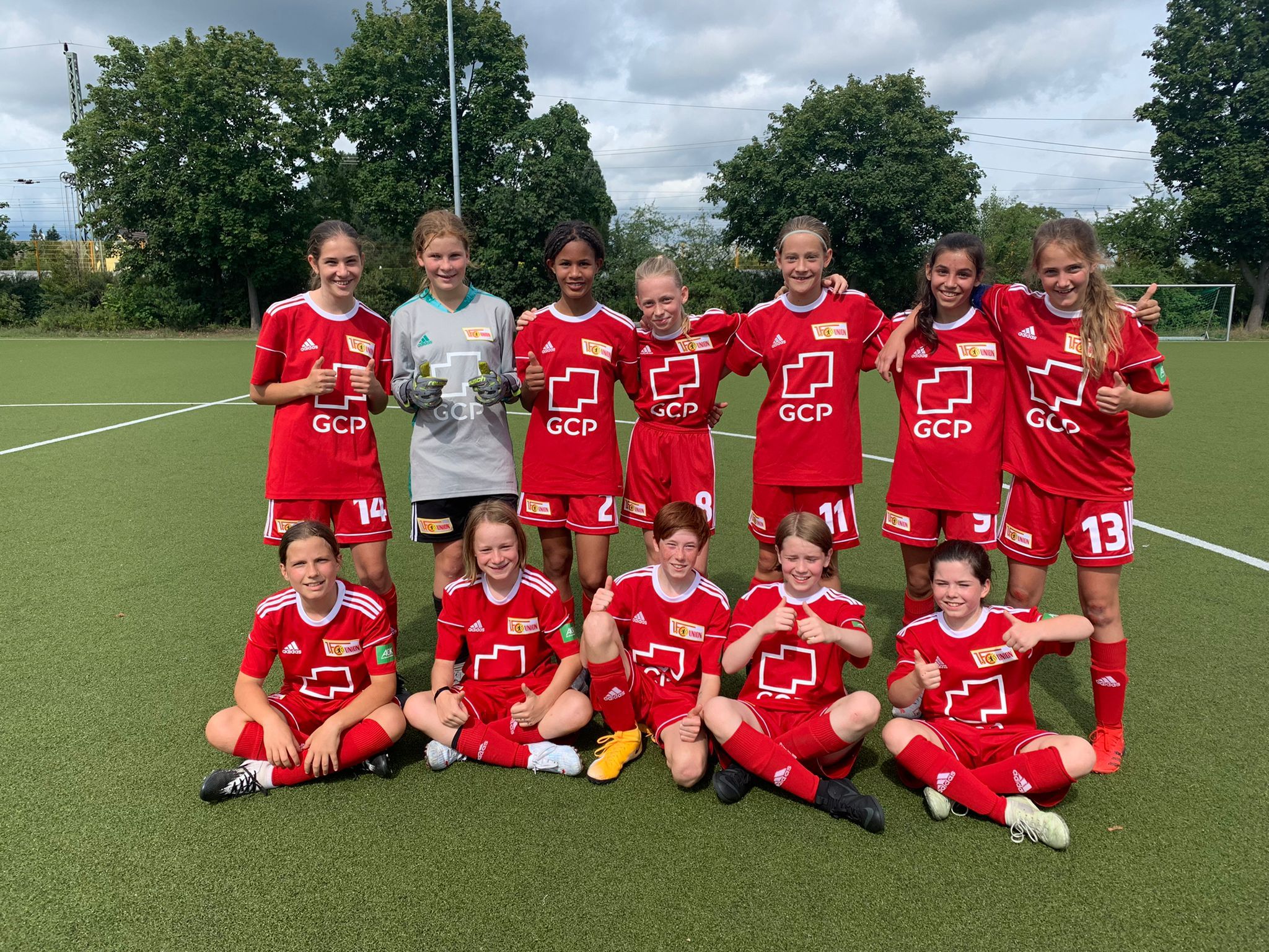 Mädchen-Fußballmannschaft in roten Trikots posiert auf einem Sportplatz, einige zeigen Daumen nach oben.
