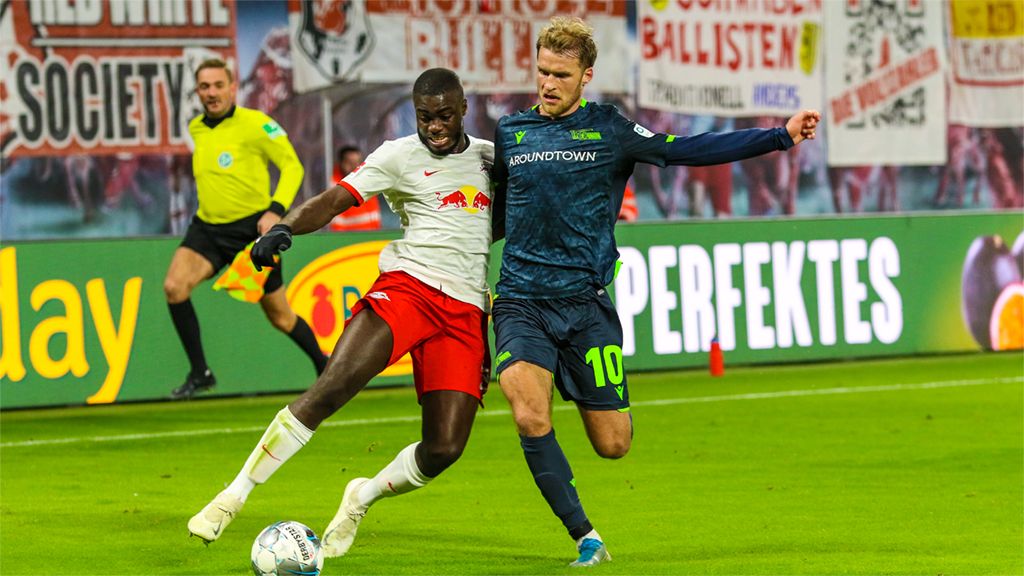 A soccer player in a red jersey is trying to win the ball from a player in a blue jersey, while fans cheer in the background.