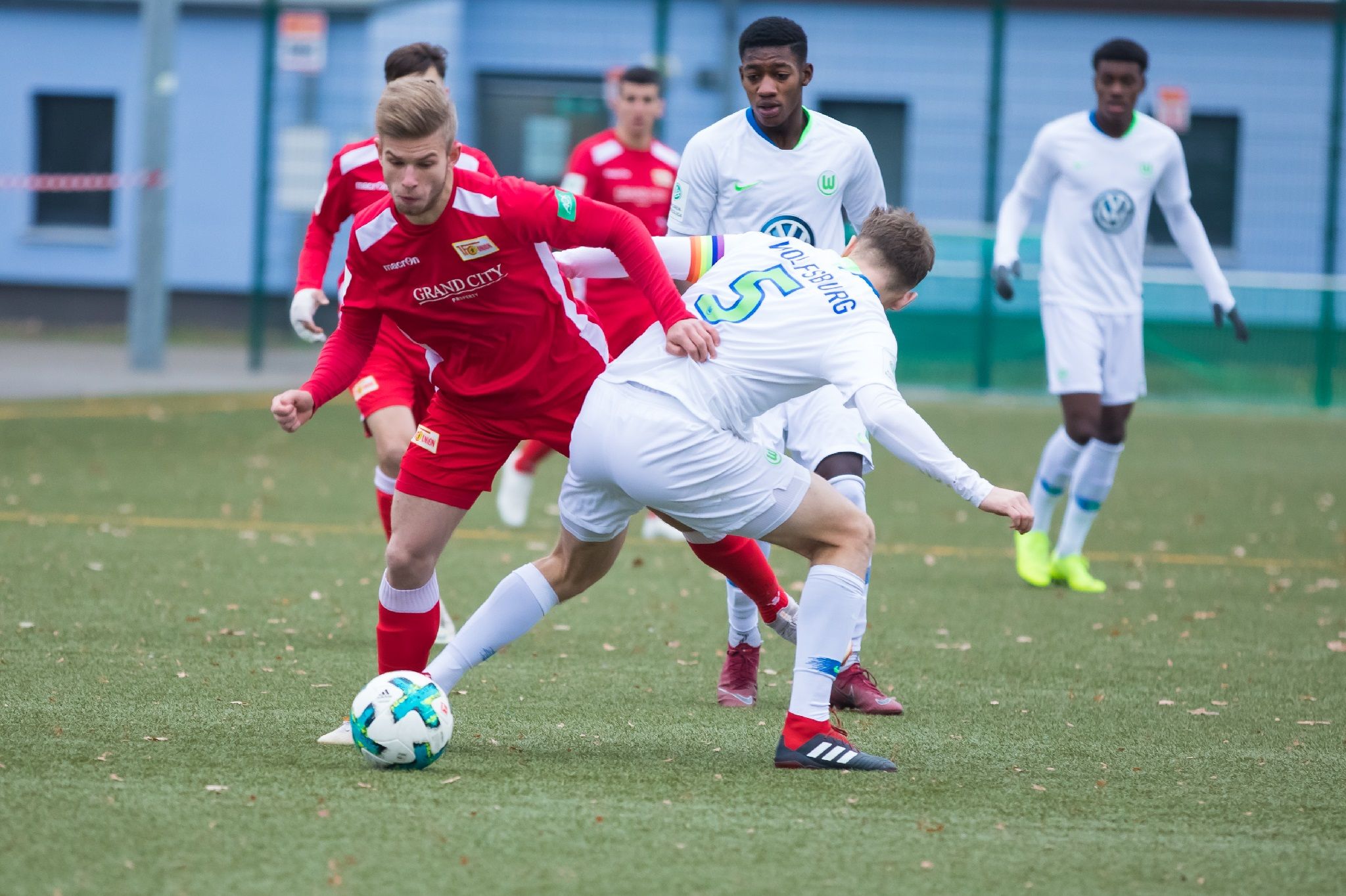 Two soccer players are fighting for the ball on an artificial turf field, while other players stand in the background.