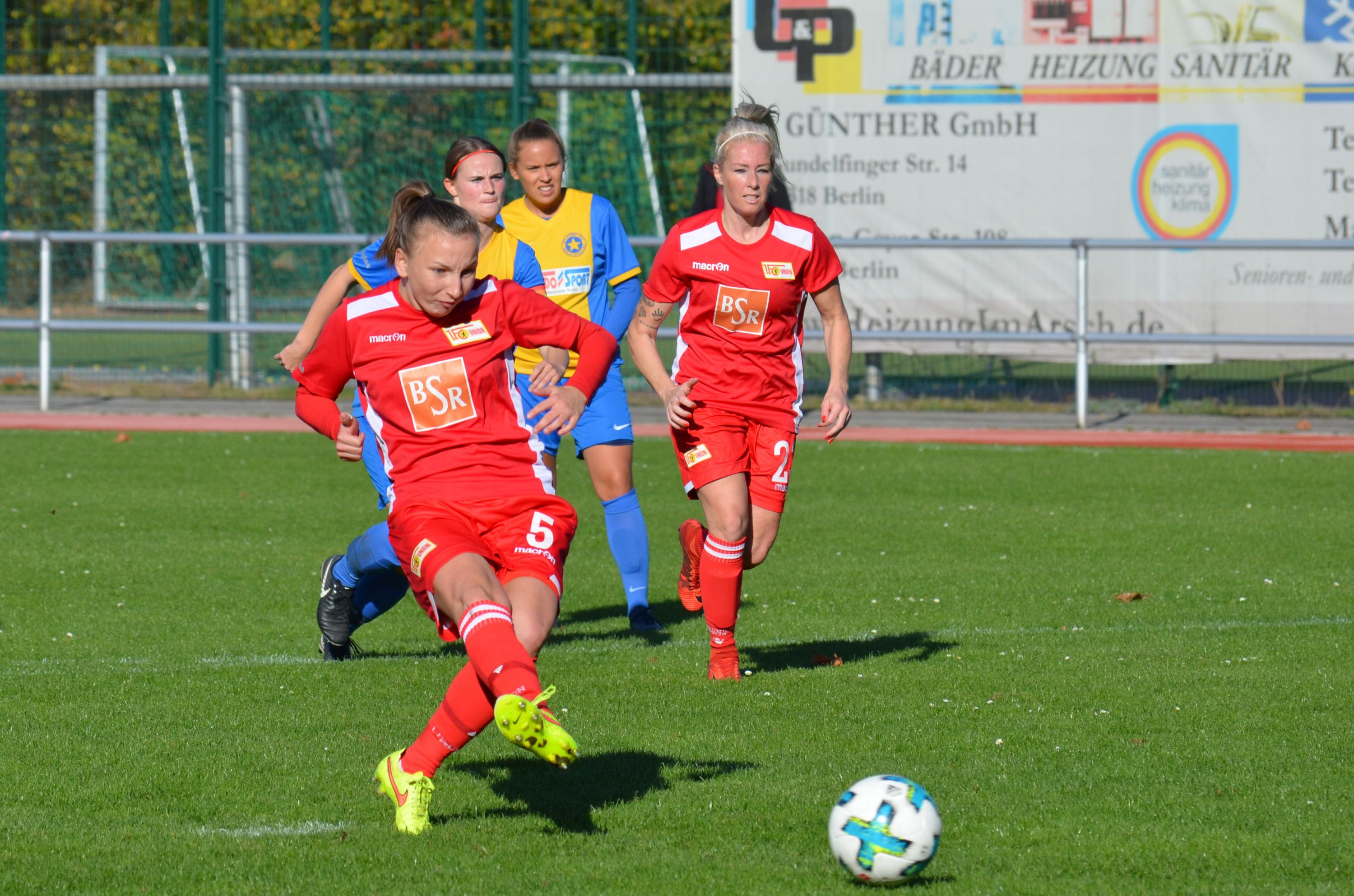 Eine Fußballerin trägt ein rotes Trikot. Sie tritt den Ball. Das Spiel ist auf einem grünen Platz.