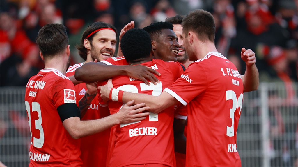 Die Fußballspieler haben ein Tor geschossen. Sie feiern und umarmen sich. Im Hintergrund sind die Fans zu sehen.