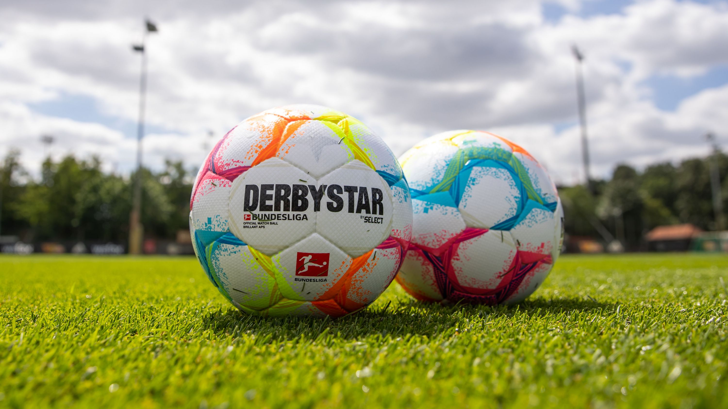 Two colorful soccer balls with the print "DERBYSTAR" on a green grass field under a cloudy sky.