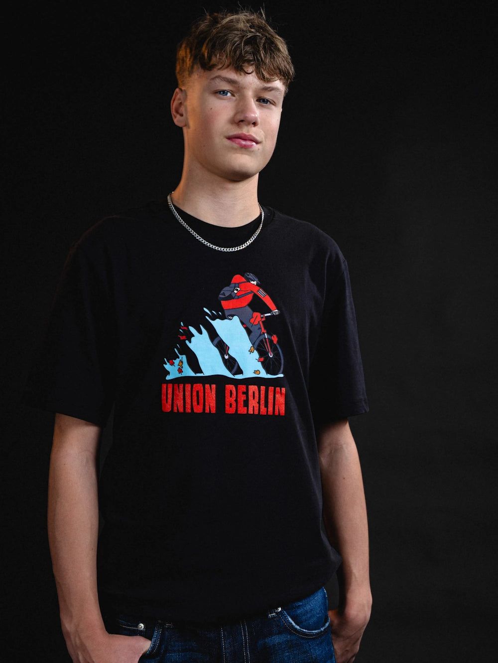 Junger Mann in schwarzem T-Shirt mit dem Aufdruck „UNION BERLIN“ vor einem dunklen Hintergrund.