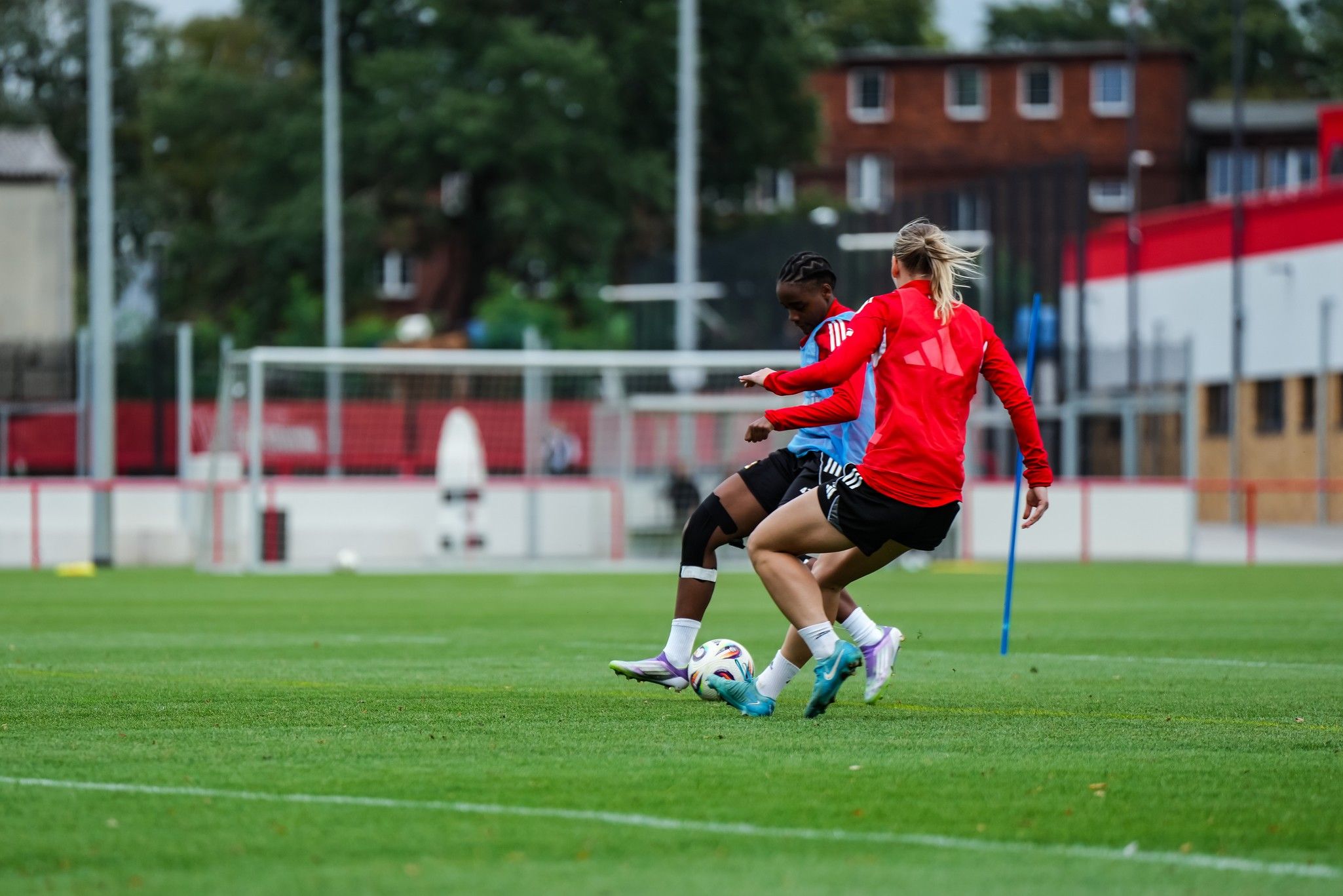 Zwei Fußballspieler in Training, einer im roten, einer im blauen Trikot, kämpfen um den Ball auf einem grünen Platz.