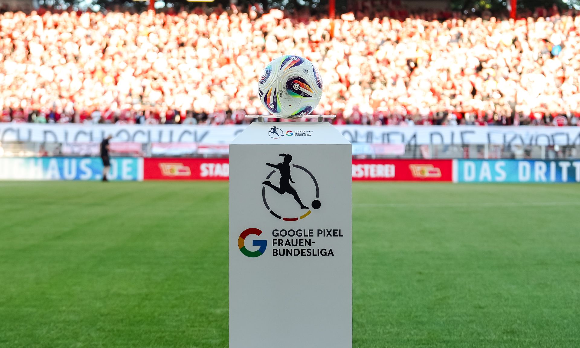 Ein Fußball steht auf einem Podest mit dem Logo der Google Pixel Frauen-Bundesliga, im Hintergrund steht eine jubelnde Menge.