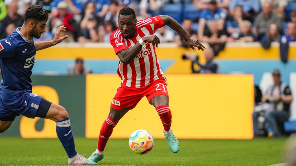 Ein Spieler mit rotem Trikot dribbelt den Ball. Ein blauer Spieler verteidigt. Im Hintergrund ist ein Stadion.