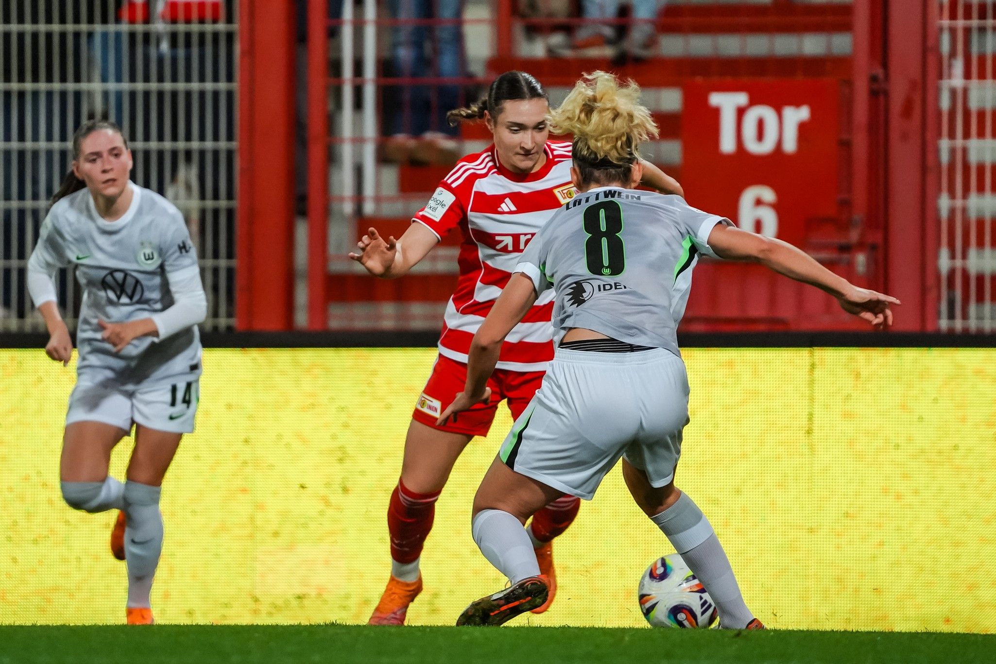 Zwei Frauenfußballspielerinnen kämpfen am Ball, während eine dritte Spielerin im Hintergrund steht. Stadionbeleuchtung im Hintergrund.