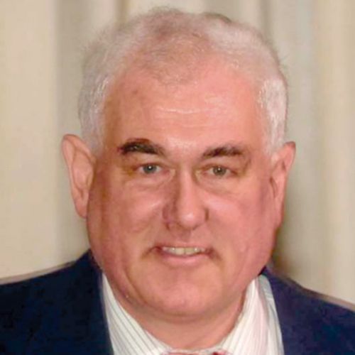 Ein Mann mit grauen Haaren und einem freundlichen Lächeln trägt einen Anzug und ist vor einem neutralen Hintergrund abgebildet.