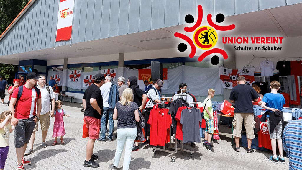 Menschen stehen an einem Stand mit Merchandising-Artikeln, umgeben von rot-weißen Flaggen und einem Logo.