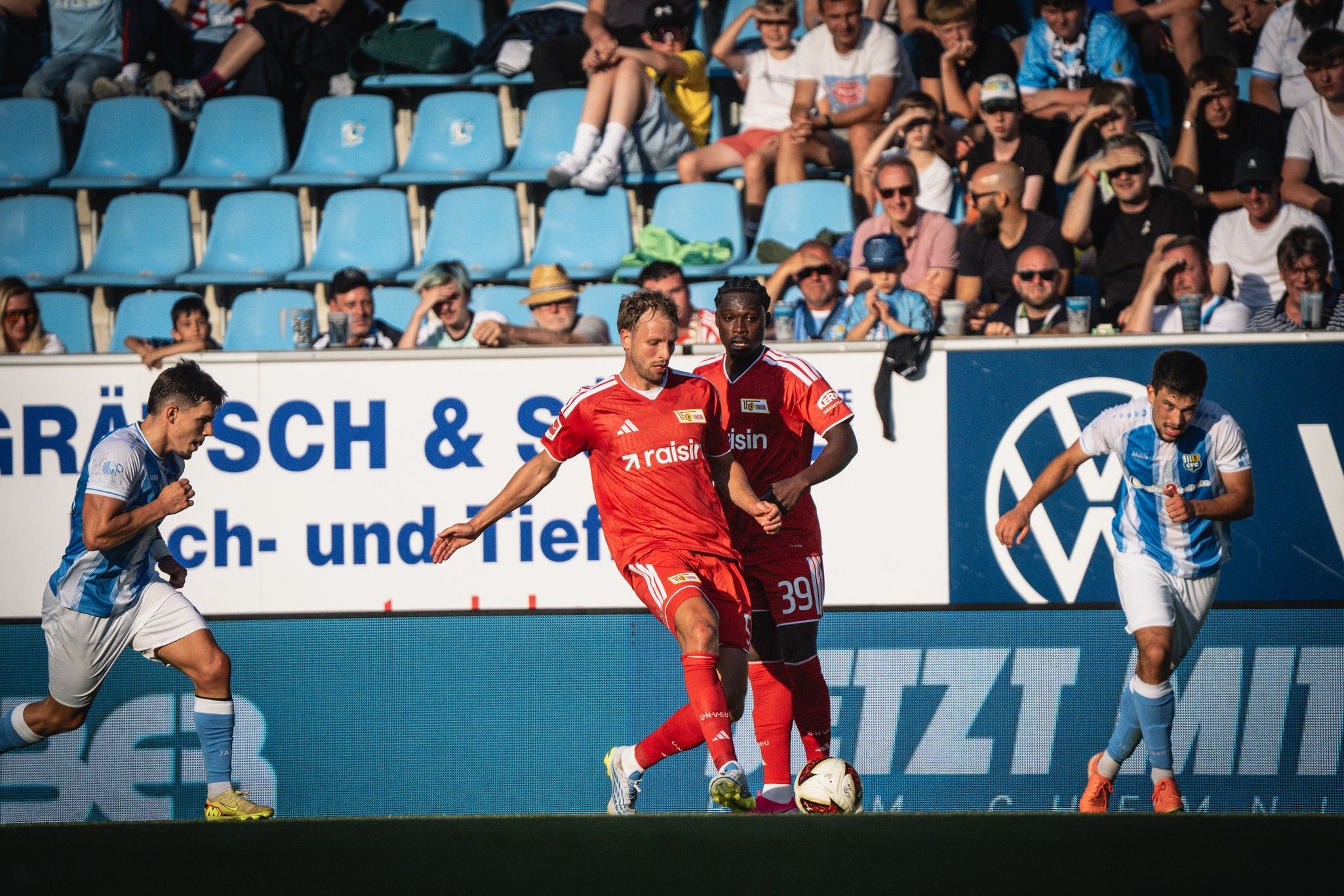 Fußballspiel mit zwei Mannschaften in roten und blauen Trikots. Ein Spieler im roten Trikot dribbelt den Ball, während Fans zuschauen.