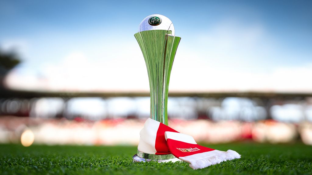 A shiny trophy stands on green grass, surrounded by red and white scarves.