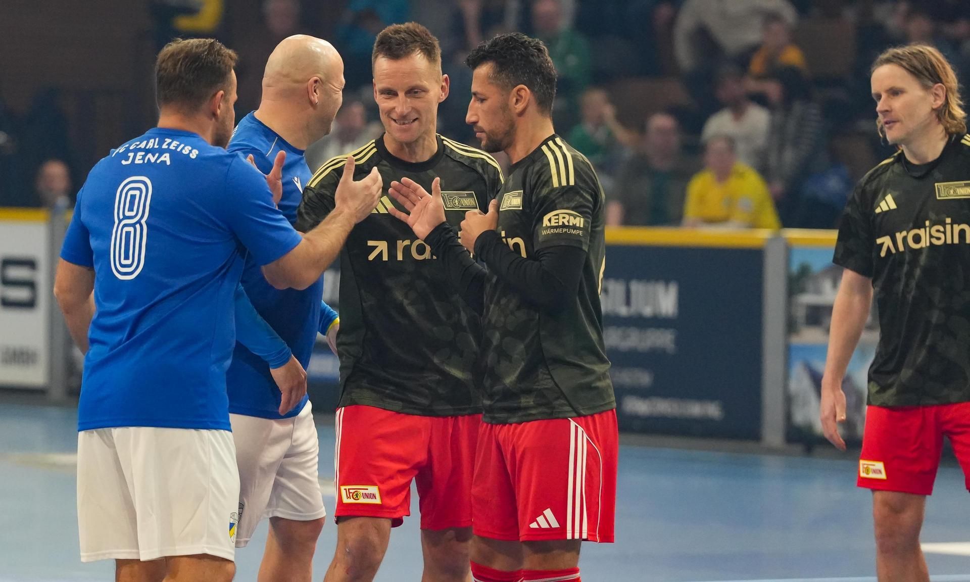 Vier Männer in Sportkleidung stehen auf einem Handballfeld, lächeln und kommunizieren miteinander.