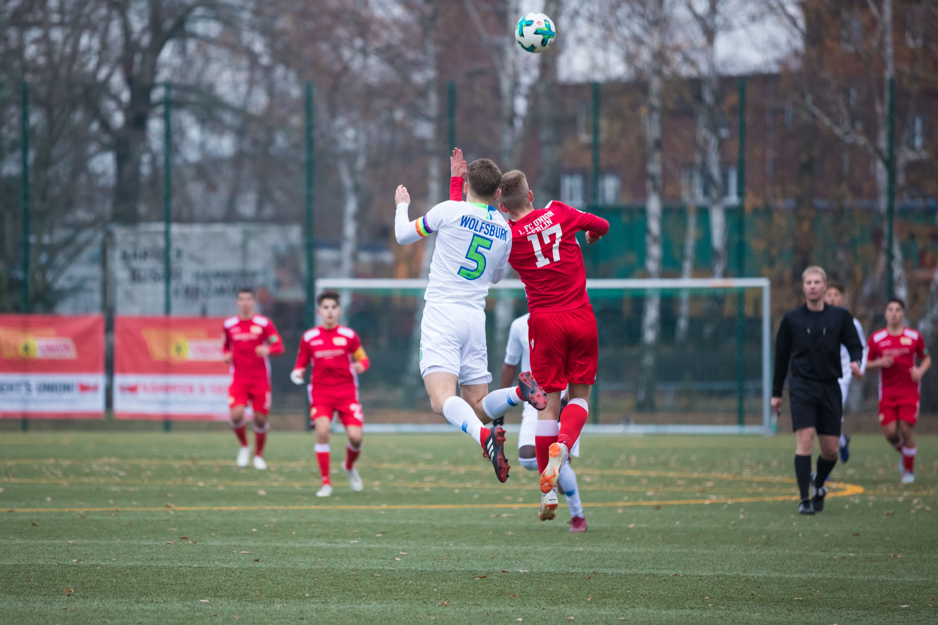 Two football players jump high to head a ball, while other players stand in the background. It is a gray, cloudy day.