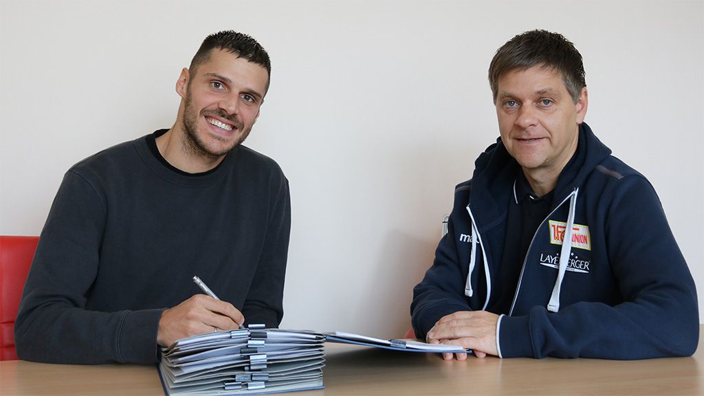 Zwei Männer sitzen an einem Tisch, einer unterschreibt Dokumente, der andere beobachtet lächelnd.