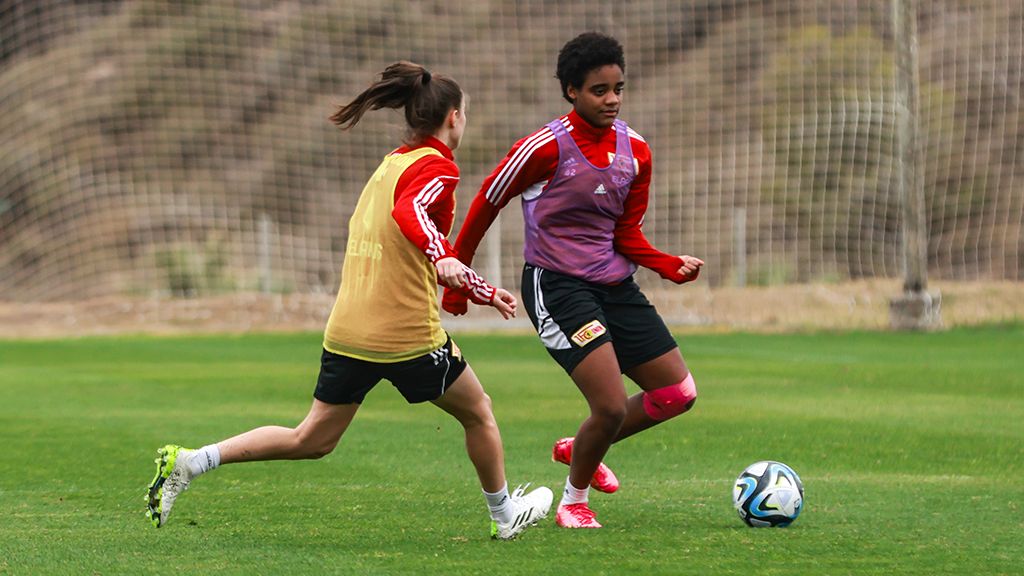 Two female soccer players are practicing on a green field, one with the ball, the other chasing her.
