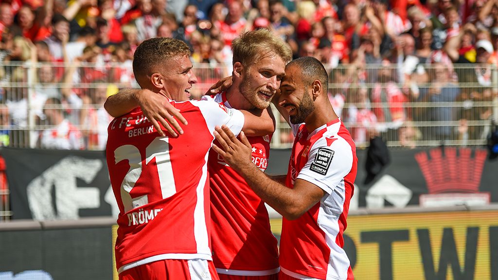 Drei Fußballspieler in roten Trikots feiern. Sie haben ein Tor geschossen. Die Fans im Stadion freuen sich.