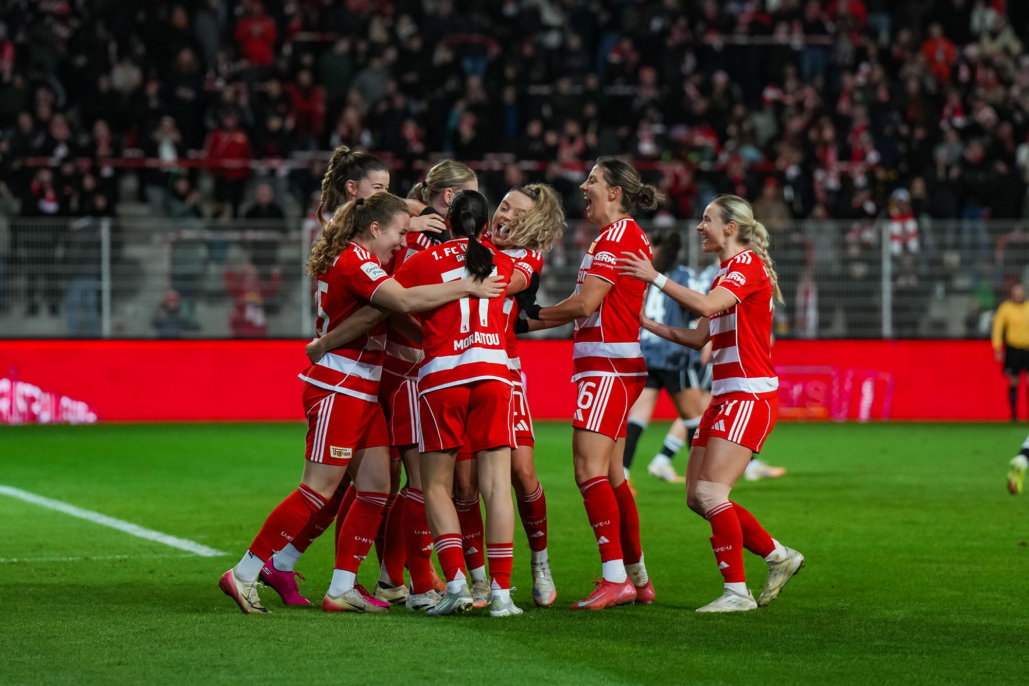 Spielerinnen eines Fußballteams in Rot feiern einen Torerfolg und umarmen sich auf dem Spielfeld. Fans sind im Hintergrund sichtbar.