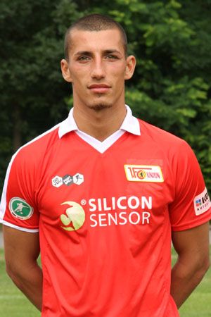 Ein junger Mann in einem roten Fußballtrikot steht vor einer grünen Hintergrundlandschaft und schaut in die Kamera.