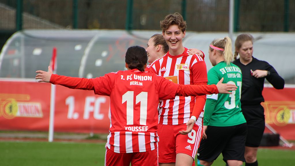 Zwei Fußballspielerinnen in roten Trikots feiern ein Tor, während eine Torhüterin im Hintergrund steht.
