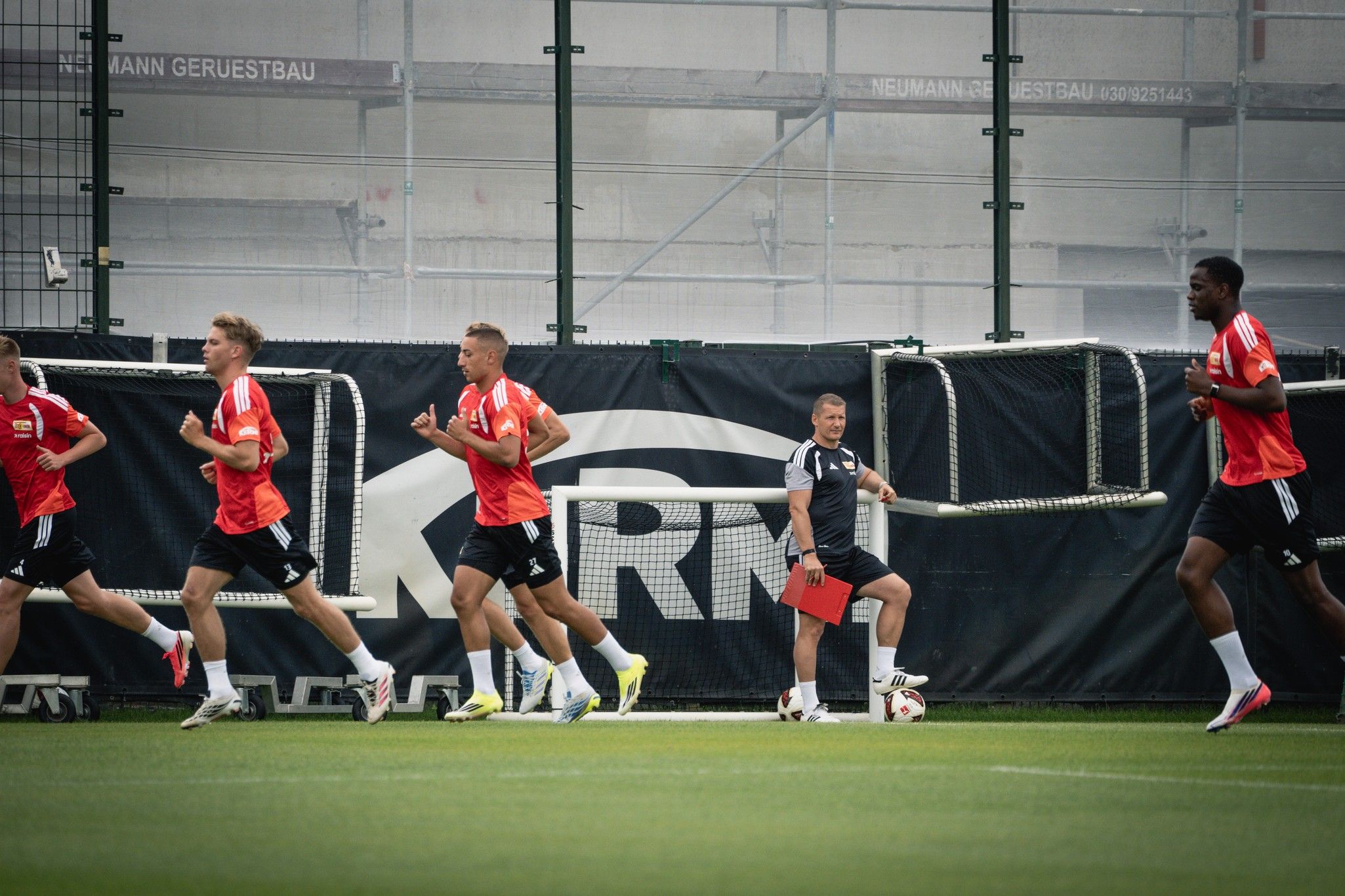Fußballspieler in roten Trikots laufen auf einem Trainingsplatz, während ein Trainer neben ihnen steht.