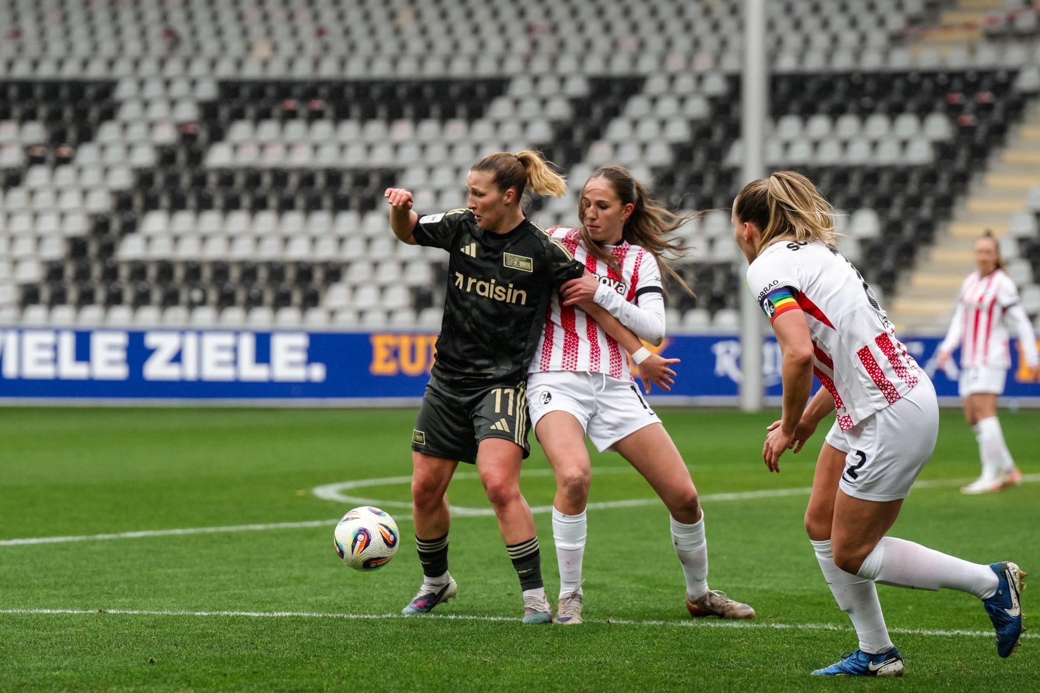 Zwei Spielerinnen im Wettkampf um den Ball, während eine dritte Spielerin im Hintergrund steht. Fußballspiel auf einem Stadionrasen.